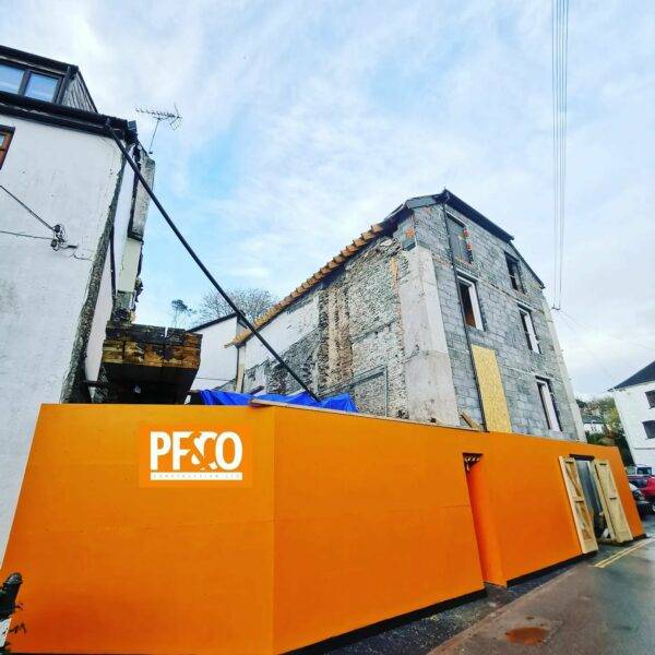 Construction project by PF & Co Construction Ltd in Cornwall, featuring a derelict building undergoing a complete rebuild with a vibrant orange site barrier in the foreground.