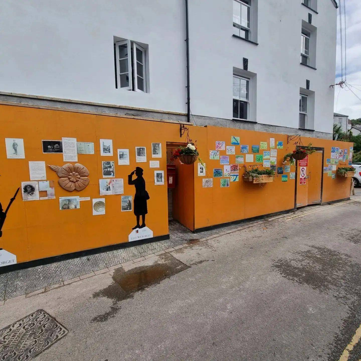 Community Remembrance Sunday display created by PF & Co Construction Ltd, featuring artwork, silhouettes, and flowers on a vibrant orange site hoarding